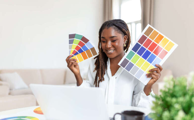 Cheerful african american lady designer having video conference with clients, sitting at desk in front of computer, holding color palettes, gesturing and smiling, copy space